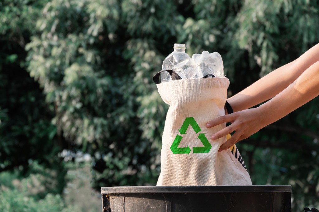 Hands holding a tote bag filled with plastic bottles, marked with a green recycling symbol